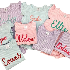 May include: A collection of eight striped shirts with personalized names embroidered on them. The shirts are in various colors, including pink, light blue, and purple. The names are embroidered in different colors, including red, green, and white.