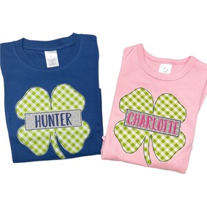 Peut inclure: Deux t-shirts avec un trèfle en gingham vert et blanc. Un t-shirt est bleu avec le nom "Hunter" sur le trèfle. L'autre t-shirt est rose avec le nom "Charlotte" sur le trèfle.