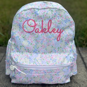 May include: A floral print backpack with the name "Oakley" embroidered in pink script. The backpack features a white zipper and is covered in a pattern of small blue, pink, and green flowers on a white background.