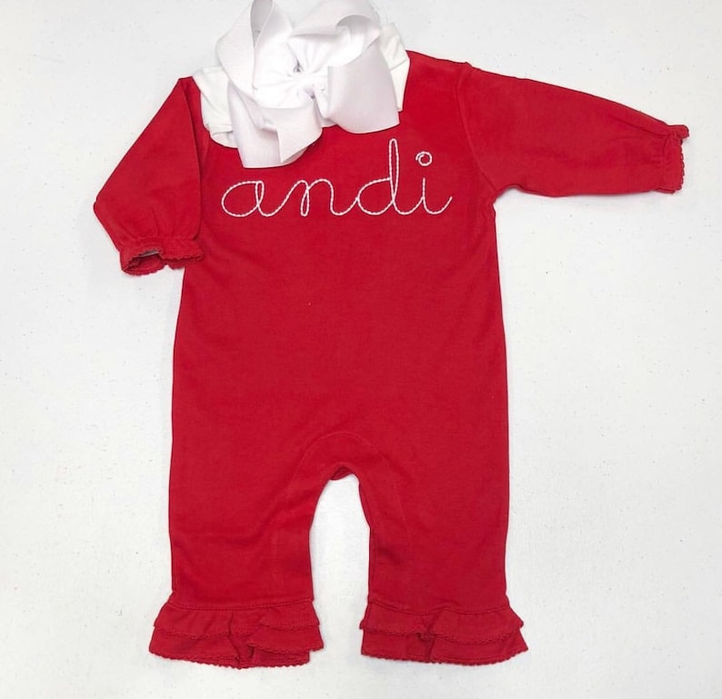 May include: Red long-sleeve baby onesie with white ruffled trim and a white bow. The onesie has the name "andi" embroidered on the front in white thread.