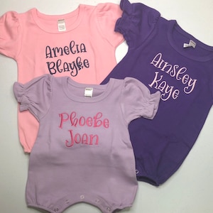 May include: Three baby onesies in pink, purple, and lavender with embroidered names. The pink onesie says "Amelia Blayke", the purple onesie says "Ainsley Kaye", and the lavender onesie says "Phoebe Joan".