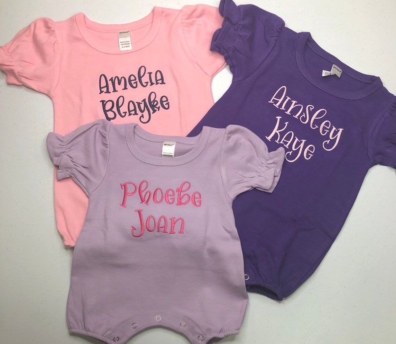 May include: Three baby onesies in pink, purple, and lavender with embroidered names. The pink onesie says "Amelia Blayke", the purple onesie says "Ainsley Kaye", and the lavender onesie says "Phoebe Joan".
