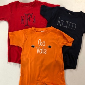 May include: Three short-sleeved t-shirts, one red with the monogram "KFJ", one navy blue with the name "Kam" and one orange with the text "Go Vols" and two footballs.