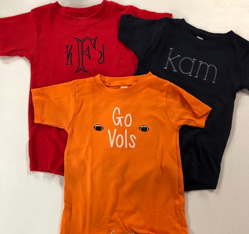 May include: Three short-sleeved t-shirts, one red with the monogram "KFJ", one navy blue with the name "Kam" and one orange with the text "Go Vols" and two footballs.