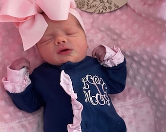 May include: A newborn baby wearing a navy blue onesie with pink ruffles and a white monogram. The baby is wearing a pink bow headband and is sleeping on a pink blanket. A wooden sign with the text "Linley-Mae Grace" is visible in the background.
