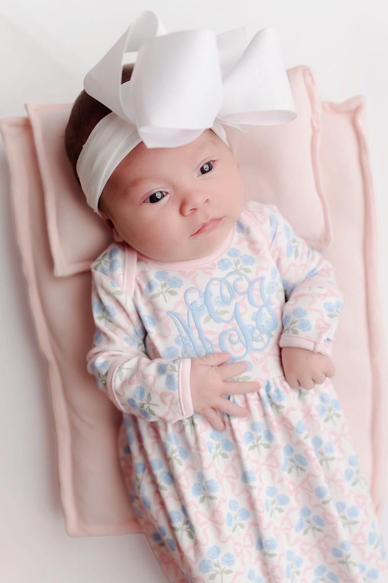 Pu&ograve; includere: Un bambino con una fascia bianca e un grande fiocco bianco. Il bambino &egrave; sdraiato su una coperta rosa e indossa una tutina a maniche lunghe con motivo floreale e monogramma azzurro. La tutina ha finiture rosa.