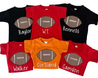 May include: A collection of short-sleeved t-shirts in black, red, and orange. Each shirt features a football design with a brown and white pattern. Names like "Baylor", "W.T.", "Bennett", "Walker", "Cortland", and "Camden" are printed on the shirts.