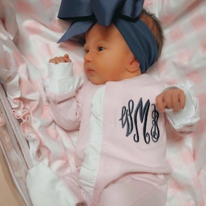 May include: A pink baby onesie with white ruffles and a navy blue monogram on the front. The baby is wearing a navy blue bow headband.