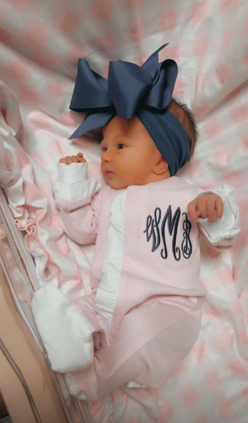May include: A pink baby onesie with white ruffles and a navy blue monogram on the front. The baby is wearing a navy blue bow headband.