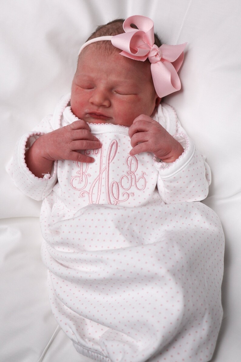 May include: A newborn baby wearing a white onesie with pink polka dots and a pink monogram. The onesie has long sleeves with scalloped edges. The baby is also wearing a pink bow headband. The baby is lying on a white surface.