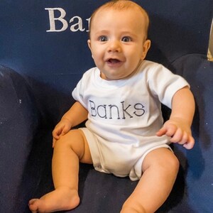 May include: A baby boy wearing a white onesie with the name "Banks" embroidered in blue on the front. The baby is sitting on a blue chair and smiling.