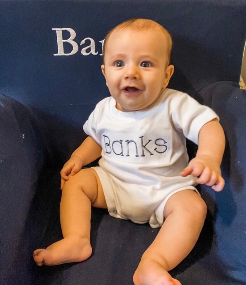 May include: A baby boy wearing a white onesie with the name "Banks" embroidered in blue on the front. The baby is sitting on a blue chair and smiling.