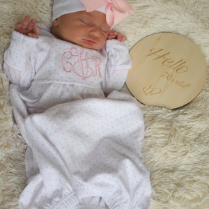 May include: A newborn baby wearing a white and pink polka dot gown and a gray hat with a pink bow. The gown has a pink monogram. A wooden disc with "Hello World!" is next to the baby.