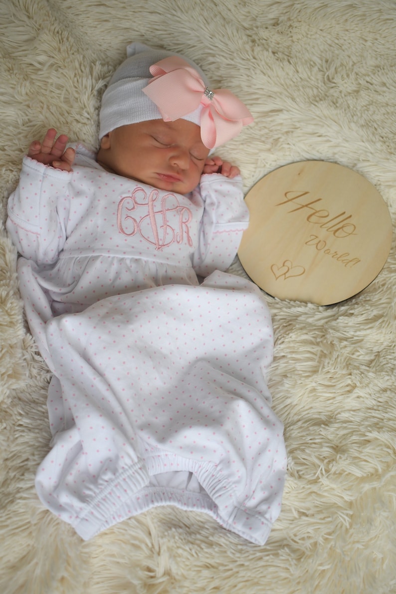 May include: A newborn baby wearing a white and pink polka dot gown and a gray hat with a pink bow. The gown has a pink monogram. A wooden disc with "Hello World!" is next to the baby.