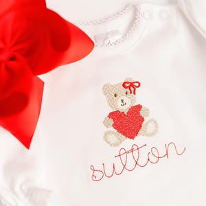 May include: White baby onesie with a red embroidered teddy bear holding a heart. The name "Sutton" is embroidered in red script below the bear. A large red bow is in the upper left corner.