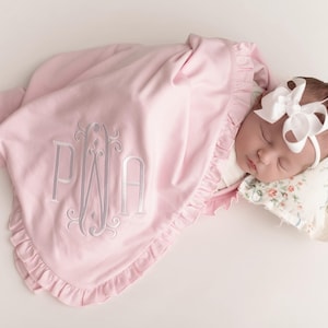 May include: A sleeping baby wrapped in a light pink blanket with a ruffled edge. The blanket features a monogram in silver thread. The baby wears a white bow headband and rests on a floral pillow.