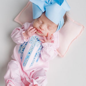 May include: A newborn baby wearing a light pink onesie with a floral monogram and ruffled trim, and a large light blue bow headband. The baby is lying on a pink pillow.
