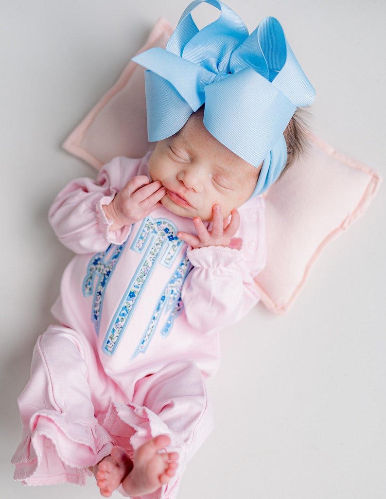 May include: A newborn baby wearing a light pink onesie with a floral monogram and ruffled trim, and a large light blue bow headband. The baby is lying on a pink pillow.