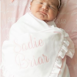 Op de afbeelding: Een wit babydekentje met roze geruite randen en de naam "Sadie Briar" geborduurd in roze. Een pasgeboren babymeisje is gewikkeld in het dekentje en draagt een roze strikband.
