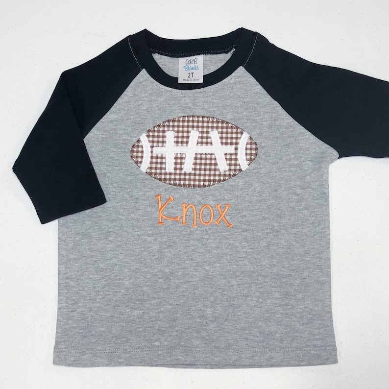 May include: A gray and black baseball tee shirt with a brown and white gingham football design and the name "Knox" embroidered in orange.