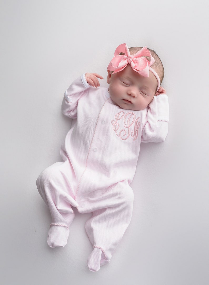 May include: A pink baby onesie with a pink embroidered monogram on the front. The onesie has long sleeves and footed bottoms. The baby is wearing a pink bow headband.