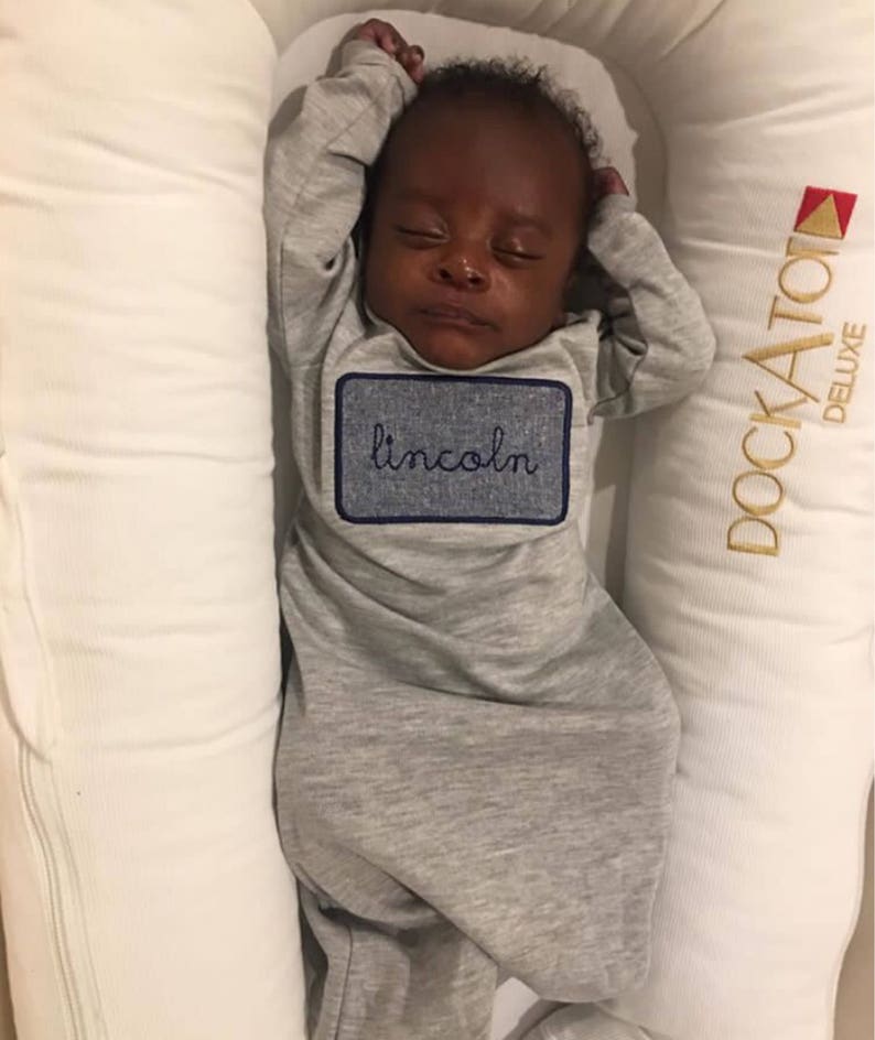May include: A sleeping baby wearing a gray onesie with a blue embroidered patch that says "lincoln". The baby is lying on a white and gold pillow with the word "docka tot" embroidered on it.