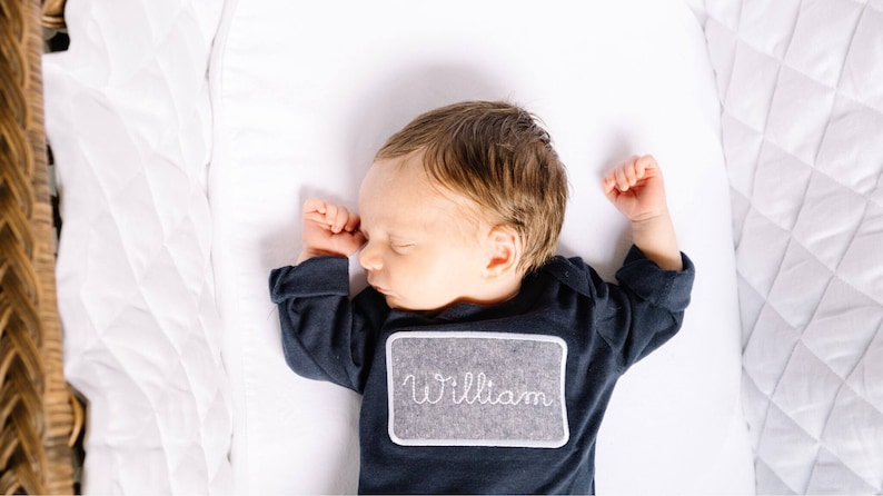 May include: A newborn baby boy sleeping in a white crib, wearing a navy blue long-sleeved onesie with a gray felt name tag that says "William".