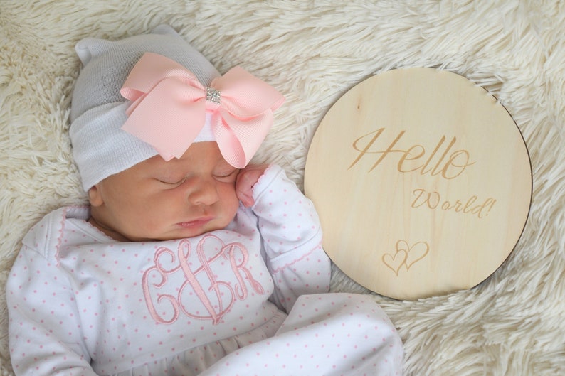 May include: A newborn baby wearing a gray hat with a pink bow and a white and pink polka dot onesie with a monogram. A wooden sign with the words "Hello World!" is next to the baby.