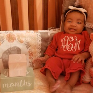 May include: A baby girl in a red outfit with white embroidered initials "H.C.M." is sitting in a crib.  She is holding a pink block with the number "4" on it.  The block is on top of a green block with the words "4 months" on it.