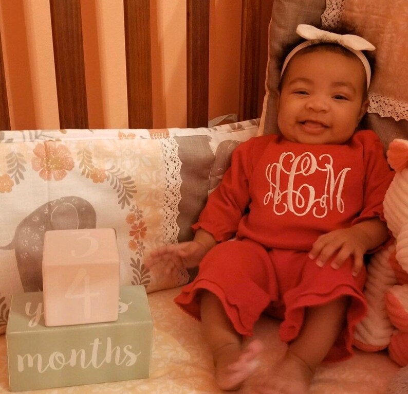 May include: A baby girl in a red outfit with white embroidered initials "H.C.M." is sitting in a crib.  She is holding a pink block with the number "4" on it.  The block is on top of a green block with the words "4 months" on it.