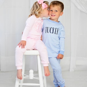Puede incluir: Una ni&ntilde;a con pijama de rayas rosas besa a un ni&ntilde;o con pijama de rayas azules. El pijama del ni&ntilde;o tiene el nombre "TUCKER" bordado en la parte delantera.