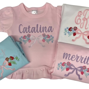May include: Children's pyjamas in pink and light blue, featuring embroidered designs. The pink top is embroidered with the name "Catalina" in purple. The pyjamas have strawberry and bow motifs. Other sets are embroidered with "Lucy", "Merritt", and initials.