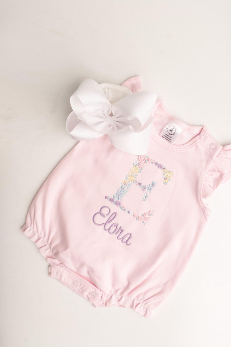 May include: A light pink baby romper with the name "Elora" embroidered in purple script. The letter "E" is decorated with floral embroidery in pastel colors. A large white bow is placed at the top.