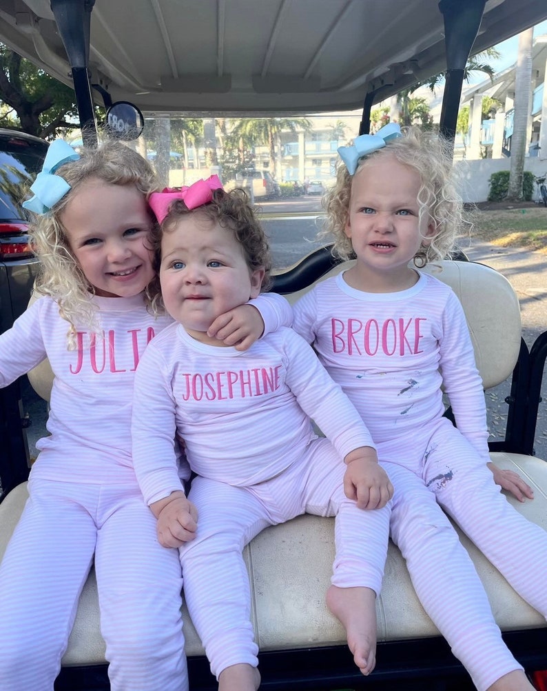 Puede incluir: Tres ni&ntilde;as peque&ntilde;as con pijamas de rayas blancas y rosas con sus nombres, "Julia", "Josephine" y "Brooke", bordados en la parte delantera. Las ni&ntilde;as est&aacute;n sentadas en un carrito de golf.