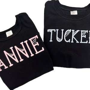 May include: Two black t-shirts with the names "ANNIE" in pink and "TUCKER" in light blue, both with a spiderweb design.