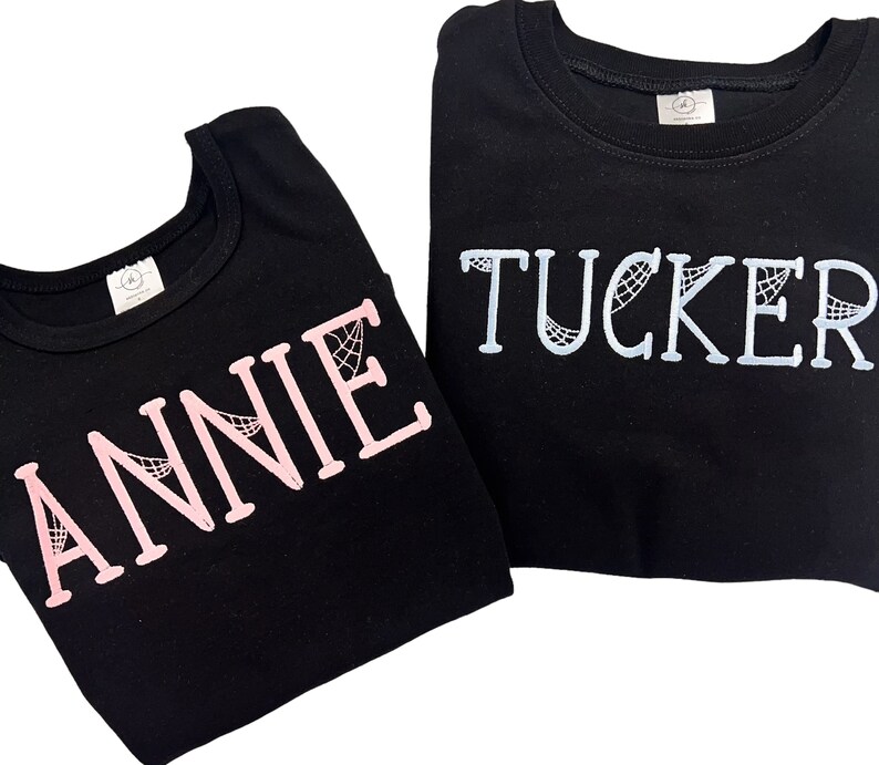 May include: Two black t-shirts with the names "ANNIE" in pink and "TUCKER" in light blue, both with a spiderweb design.