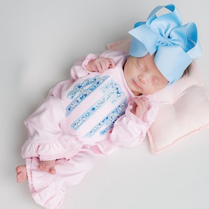 May include: A sleeping infant wearing a pink onesie with a floral design and a large blue bow. The onesie has a ruffled hem and a matching floral design on the front. The baby is lying on a small pink pillow.