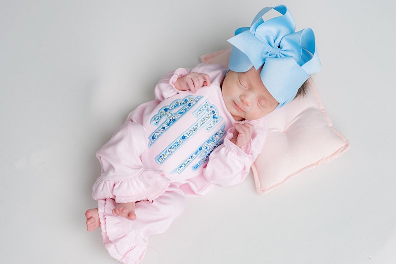 May include: A sleeping infant wearing a pink onesie with a floral design and a large blue bow. The onesie has a ruffled hem and a matching floral design on the front. The baby is lying on a small pink pillow.