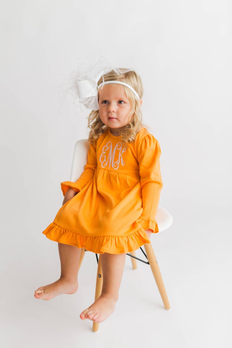 May include: A young girl wearing a bright orange dress with a white ruffle hem and a white feather headband. The dress has a white embroidered monogram "EMK" on the front.