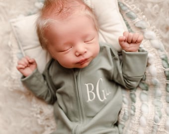 May include: A newborn baby wearing a sage green onesie with a zipper and the embroidered initials "BG". The baby is lying on a cream-colored pillow and a fluffy blanket. The baby has light hair and is sleeping.