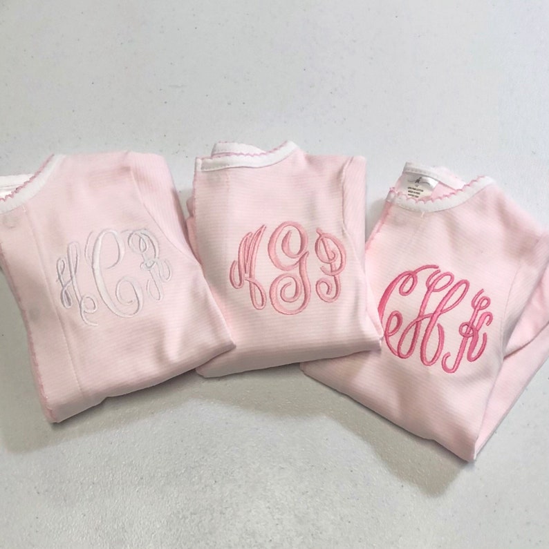 May include: Three pink long-sleeved shirts with white and pink embroidered monograms. The shirts have a white trim around the neckline.