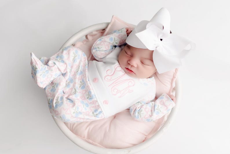 以下が含まれることがあります： 白いバスケットの中で眠っている新生児。ピンクのモノグラムと花柄のズボンが付いた白いロンパースを着ています。赤ちゃんは頭に大きな白いリボンをつけ、ピンクの枕の上に休んでいます。