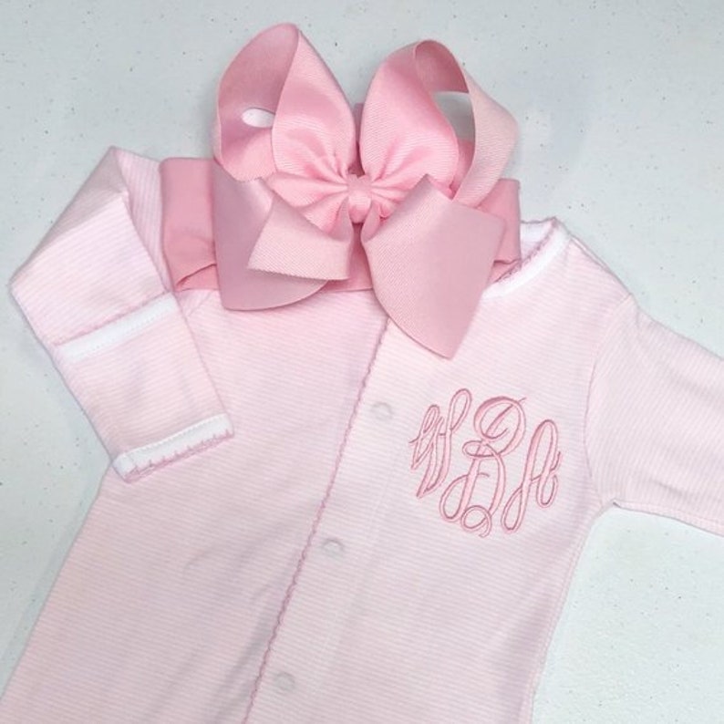 May include: Pink striped baby sleepsuit with a white trim and a pink bow on top. The sleepsuit has a monogram on the front that says "B.D.A."