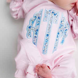 May include: A light pink baby sleeper with ruffled sleeves and pant cuffs. The sleeper features a monogram in blue floral fabric with a scalloped border. The baby's foot is visible.