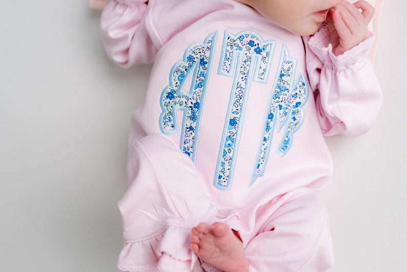May include: A light pink baby sleeper with ruffled sleeves and pant cuffs. The sleeper features a monogram in blue floral fabric with a scalloped border. The baby's foot is visible.