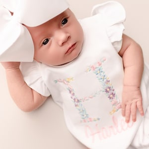 May include: A newborn baby wearing a white romper with a floral embroidered letter "H" and the name "Harley" in pink script. The baby also wears a large white bow.