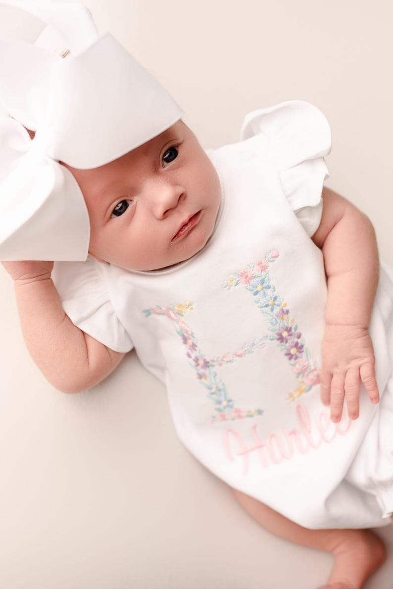 May include: A newborn baby wearing a white romper with a floral embroidered letter "H" and the name "Harley" in pink script. The baby also wears a large white bow.