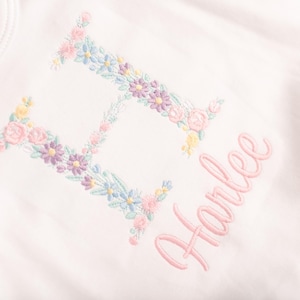 May include: White baby onesie with a floral embroidered letter "H" and the name "Harlee" in pink script. The floral embroidery includes pink, blue, yellow, and purple flowers. A tag with text is visible on the top left.