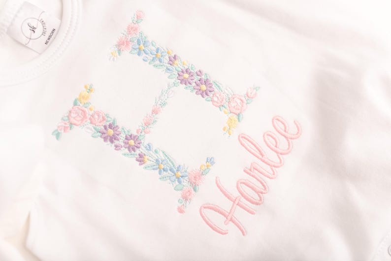May include: White baby onesie with a floral embroidered letter "H" and the name "Harlee" in pink script. The floral embroidery includes pink, blue, yellow, and purple flowers. A tag with text is visible on the top left.