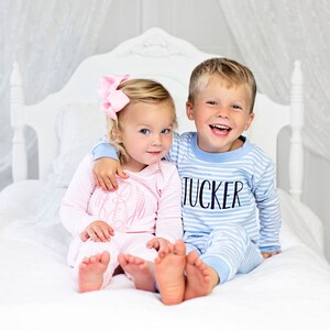 Puede incluir: Dos ni&ntilde;os con pijamas a juego est&aacute;n sentados en una cama blanca. La ni&ntilde;a lleva un pijama de rayas rosas con un lazo rosa en el pelo y el ni&ntilde;o lleva un pijama de rayas azules con el nombre "Tucker" en la parte delantera.